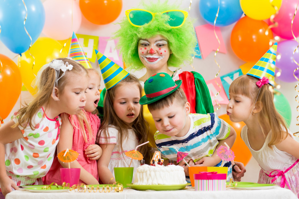 kids blowing a cake with party hats, balloons, and a clown at the back